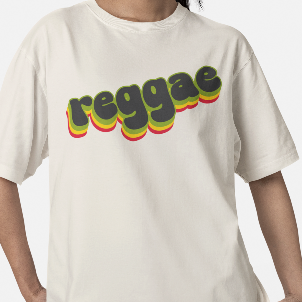 A person wearing a bone-beige short-sleeve T-shirt with the word 'reggae' printed across the chest in a retro 3D-style font with yellow, green, and red shadowing.